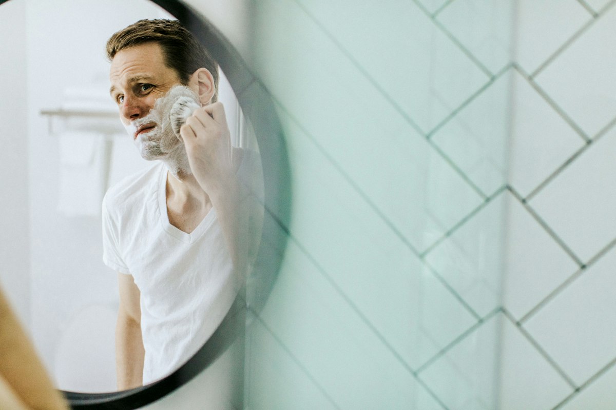 Homem a fazer a barba em frente a um espelho redondo na casa de banho