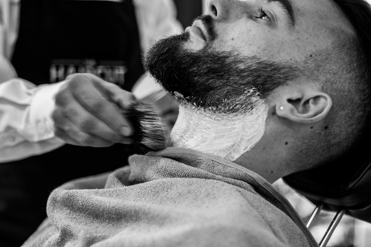 Barbeiro a aplicar espuma de barbear no pescoço de um cliente com barba, a preto e branco
