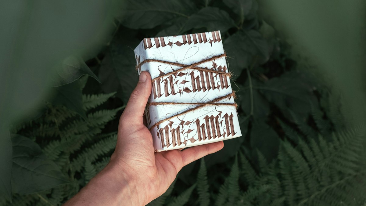 Mão a segurar um presente embrulhado em papel branco, com folhas verdes ao fundo